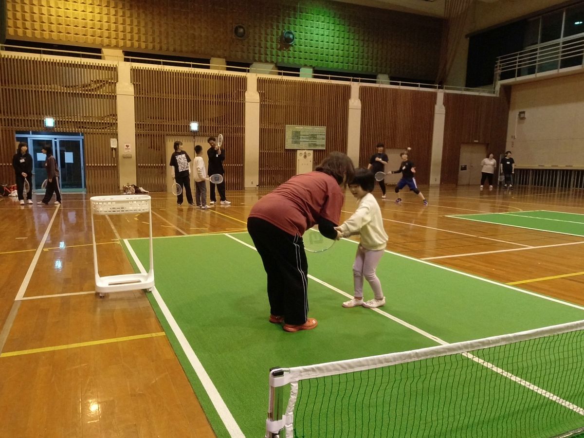 緑のコートの上で、指導者が小さな子どもにラケットの持ち方を教えているところ