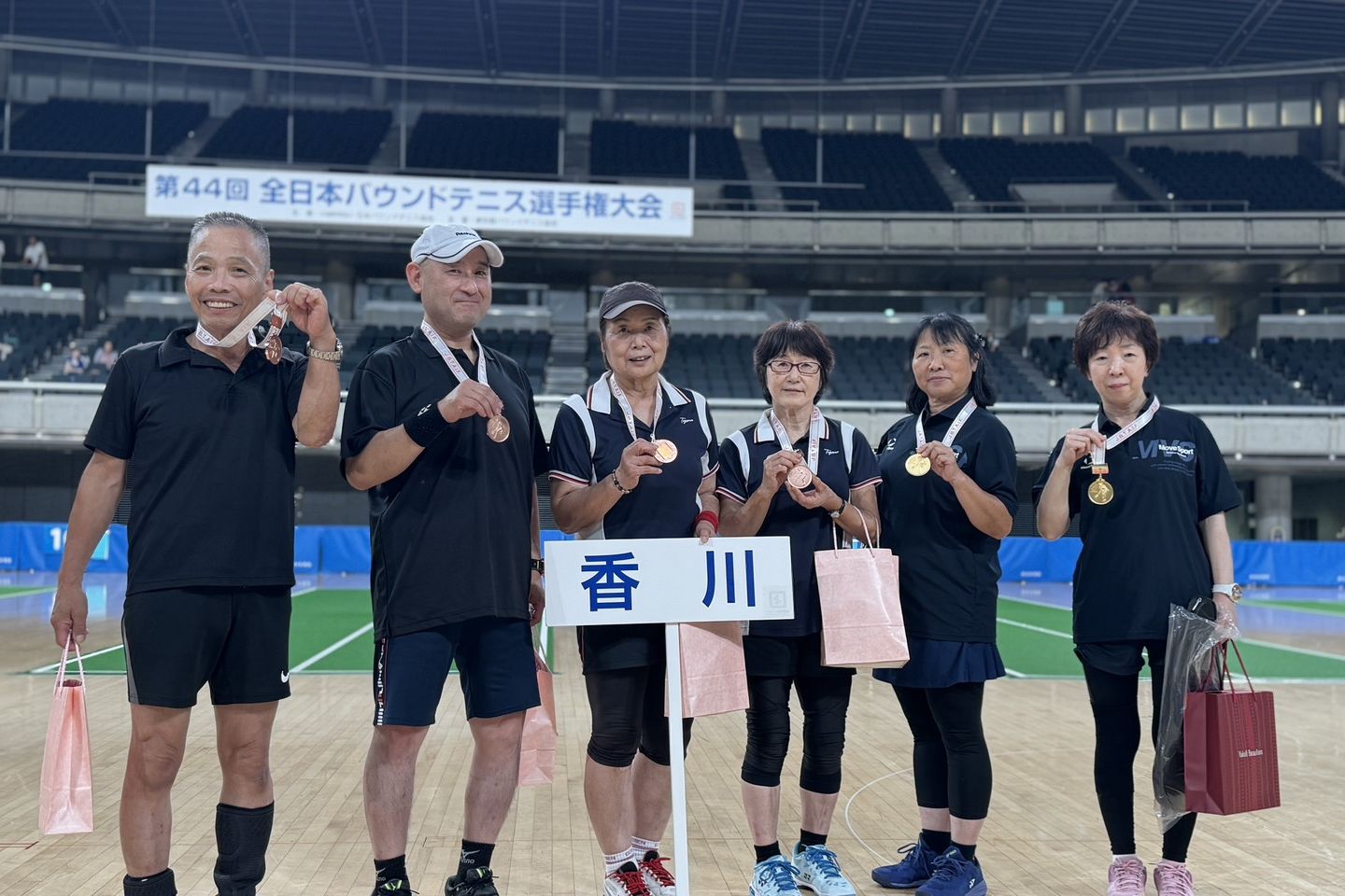 「香川」のプラカードを囲んで、首にかけたメダルを見せる香川県の選手6人。後ろに「第44回 全日本バウンドテニス選手権大会」の横断幕